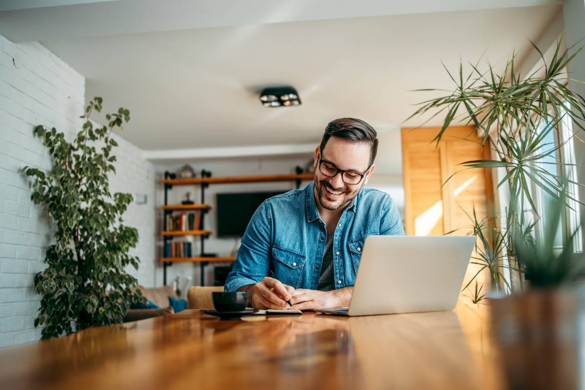 Man sitting at home with a laptop, smiling and taking notes, demonstrating the flexibility of an Online+ degree from Texas Tech University. Enjoy the convenience of learning from home while pursuing your degree on your schedule.